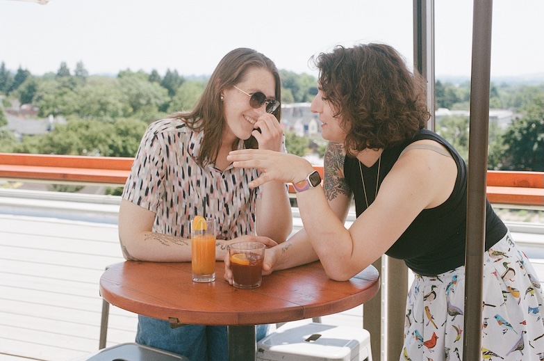 Avery and Angel standing together at a table with drinks in hand, talking